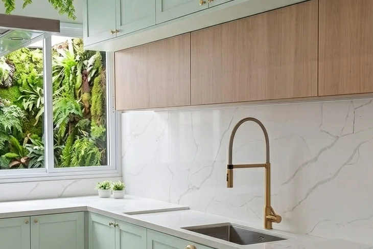 Cozinha com armários verde-menta em estilo shaker, puxadores dourados, armários superiores em madeira clara, bancada e parede em pedra branca com veios cinza, torneira gourmet dourada e janela com vista para jardim vertical